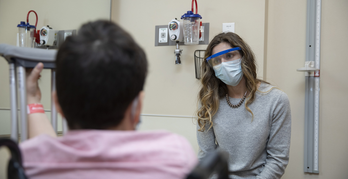 A woman wearing a face shield talks to a man in a wheelchair